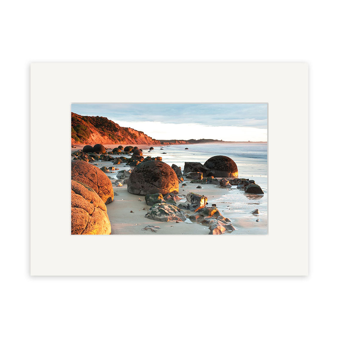 Moeraki Boulders - RP210 by Reuben Price captures large round boulders on a sandy shore, with cliffs and greenery under a partly cloudy sky, all presented in a white matte frame.
