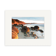 Moeraki Boulders - RP210 by Reuben Price captures large round boulders on a sandy shore, with cliffs and greenery under a partly cloudy sky, all presented in a white matte frame.