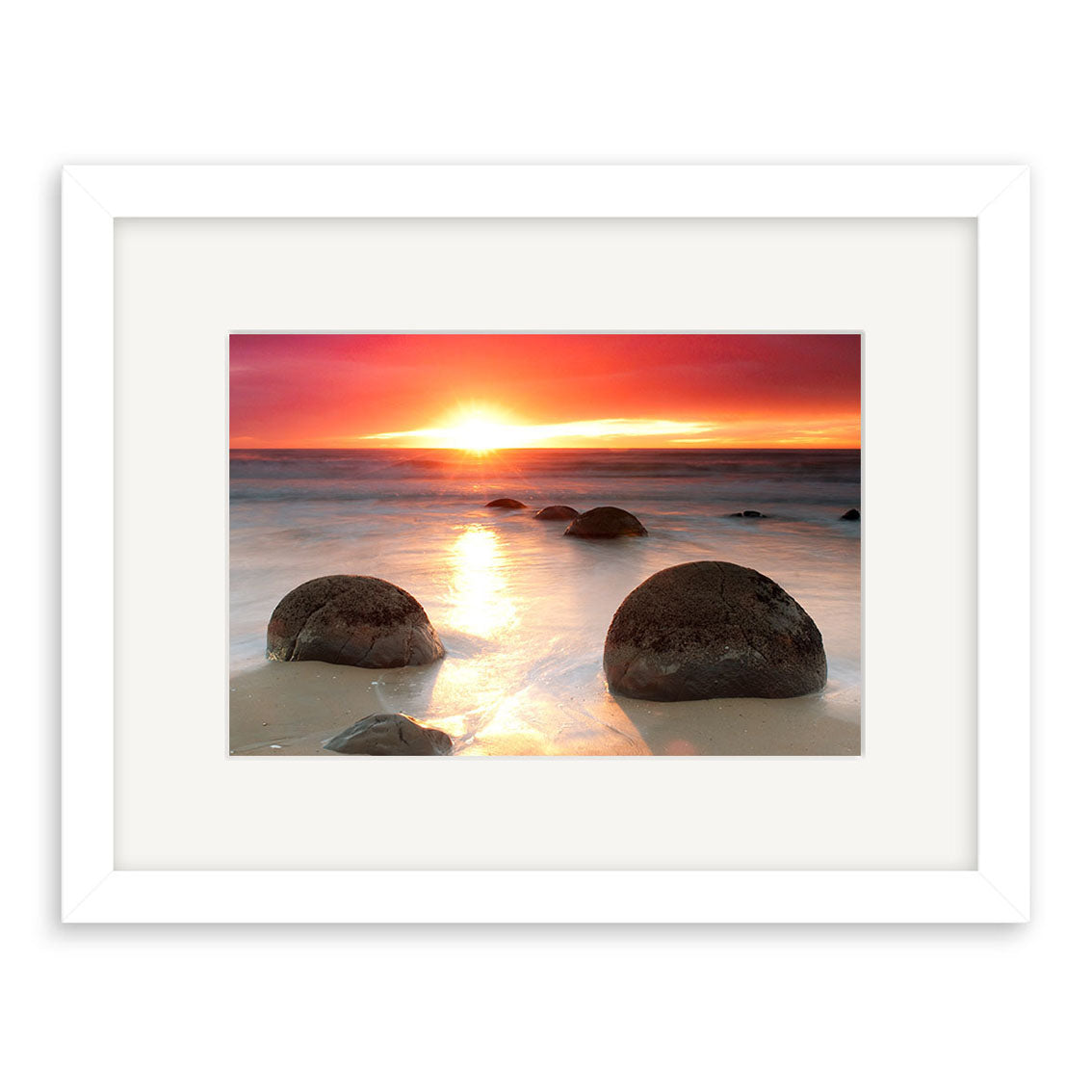 Moeraki Boulders Sunrise - RP211: A stunning photograph of round boulders on a sandy New Zealand beach at sunrise, with vibrant sky and shimmering water, displayed in a white frame from Reuben Price’s photo prints collection.