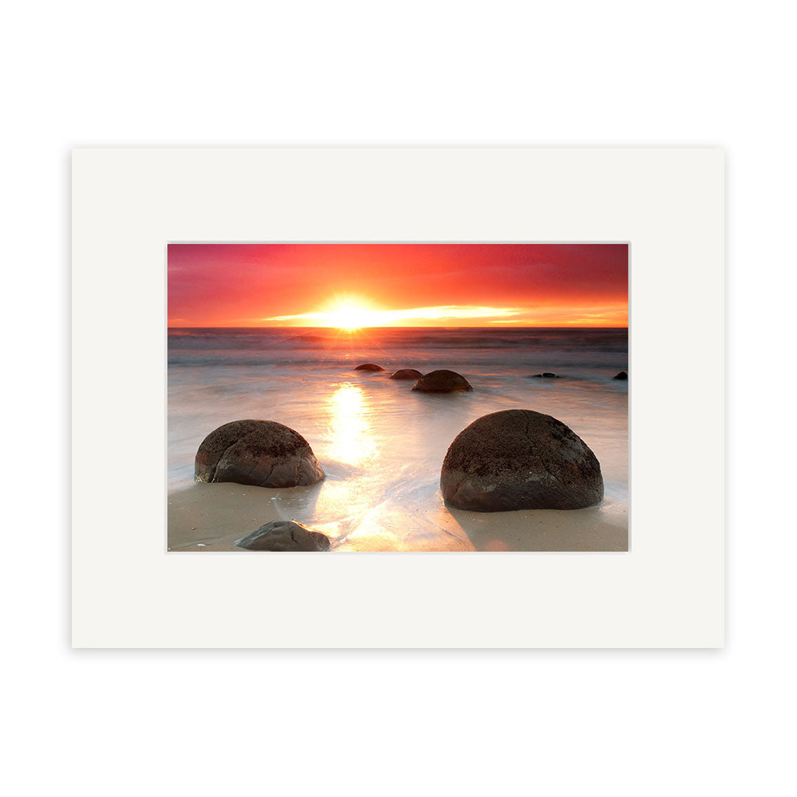 The Moeraki Boulders Sunrise - RP211 print by Reuben Price captures large round boulders on a New Zealand beach at sunrise, with vibrant orange and pink reflections and a white mat border for an elegant finish.