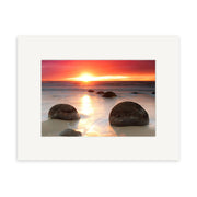 The Moeraki Boulders Sunrise - RP211 print by Reuben Price captures large round boulders on a New Zealand beach at sunrise, with vibrant orange and pink reflections and a white mat border for an elegant finish.