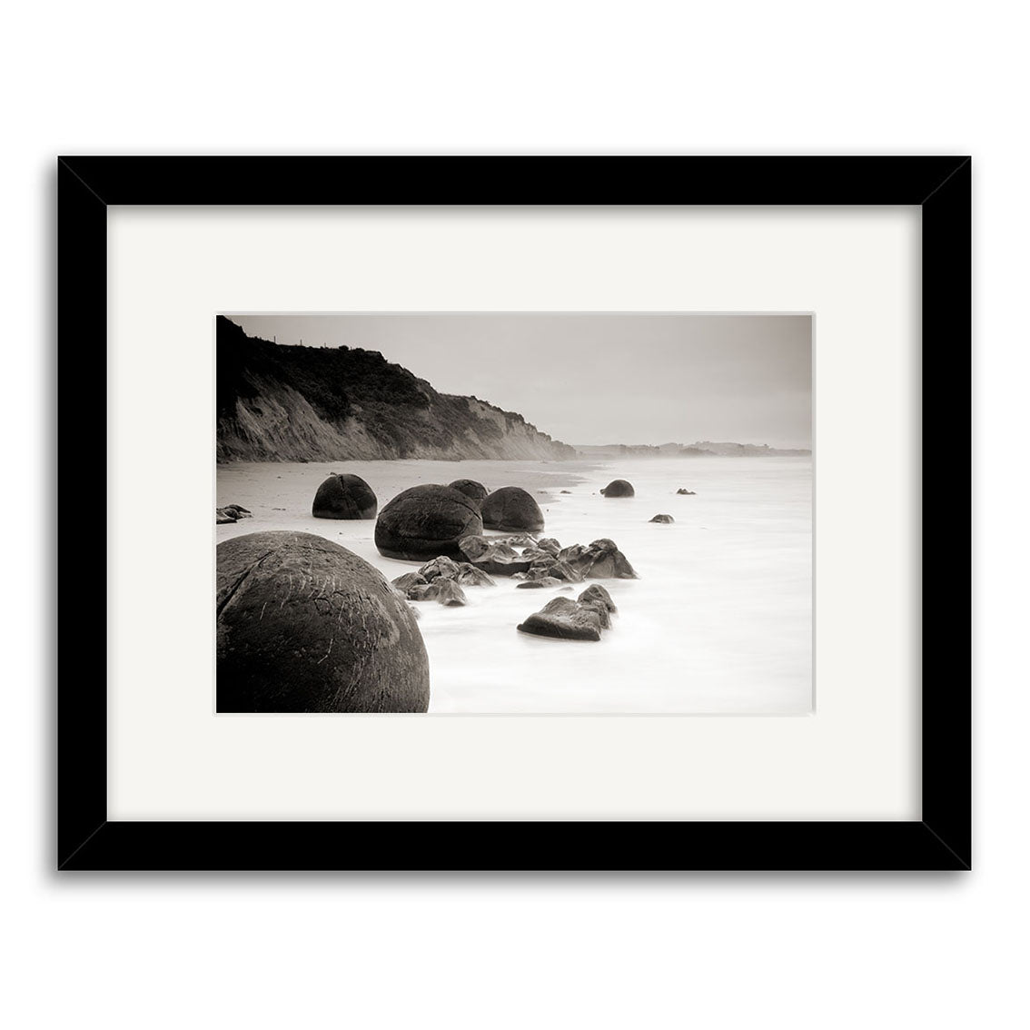 Moeraki Boulders Sepia - RP212: Sepia-toned photo of large round boulders on a misty New Zealand coast with cliffs, framed in black with a white mat and printed on archival quality matte paper.