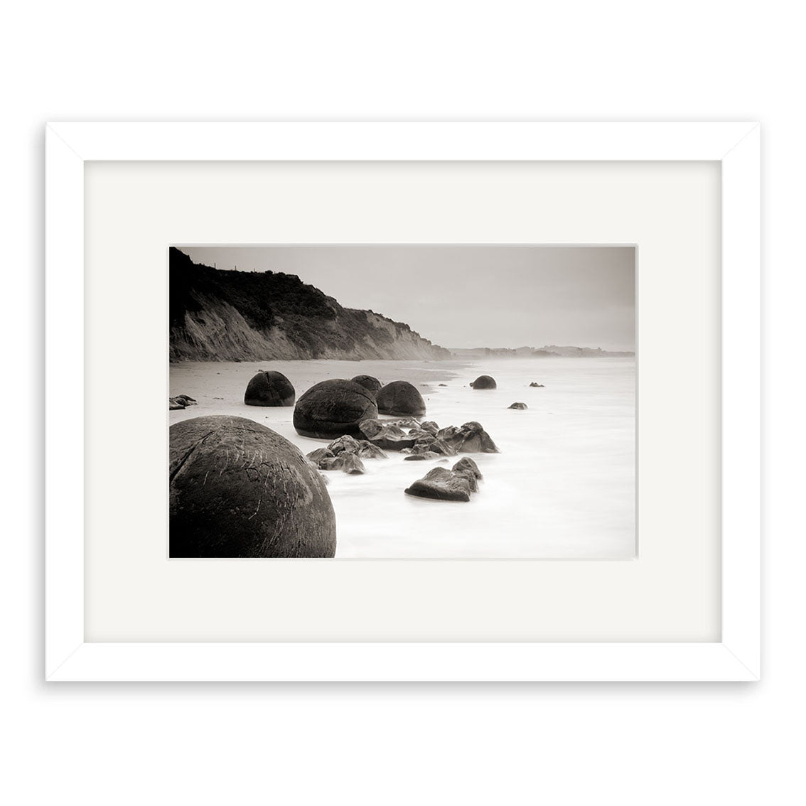 Moeraki Boulders Sepia - RP212: A framed photographic print featuring large round boulders on a misty New Zealand shore with cliffs, printed on archival matte paper and matted in white within a white frame.