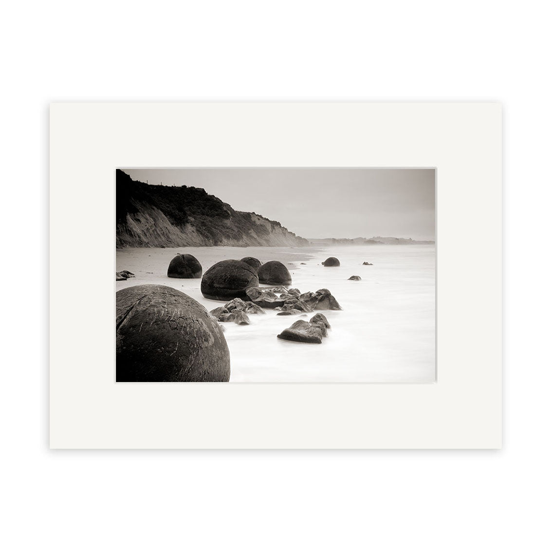 The Moeraki Boulders Sepia - RP212 print features large round boulders along New Zealand’s shore, with cliffs in the background and calm waters, beautifully framed with a white mat.