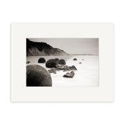 The Moeraki Boulders Sepia - RP212 print features large round boulders along New Zealand’s shore, with cliffs in the background and calm waters, beautifully framed with a white mat.