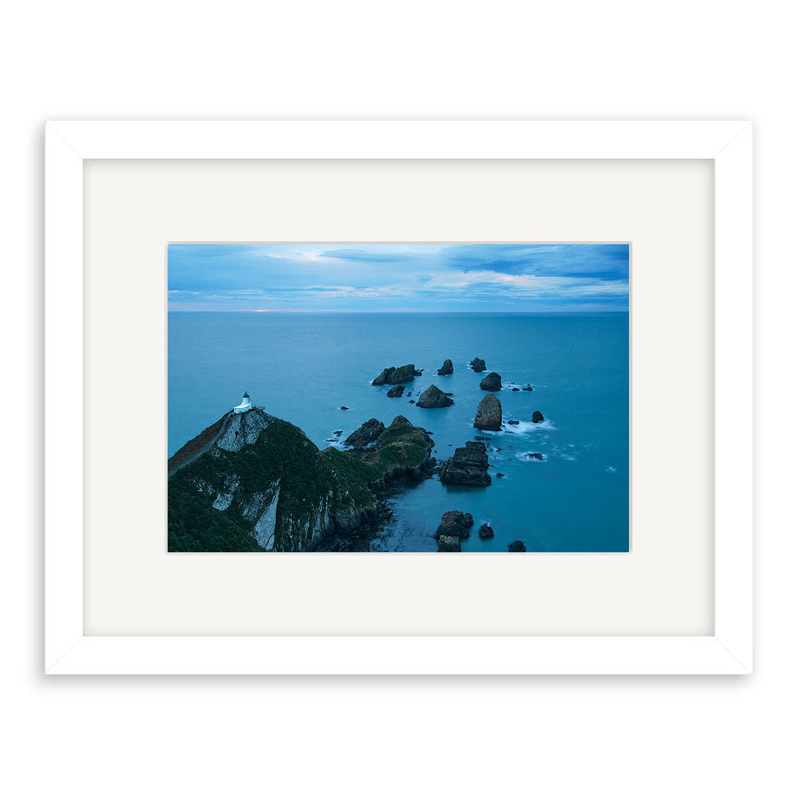 The Nugget Point Caitlins - RP214 is a framed photo of an ocean after sunset in New Zealand, with rocky formations, a green cliff, a white lighthouse, and cloudy skies. Printed on archival matte paper for lasting beauty.