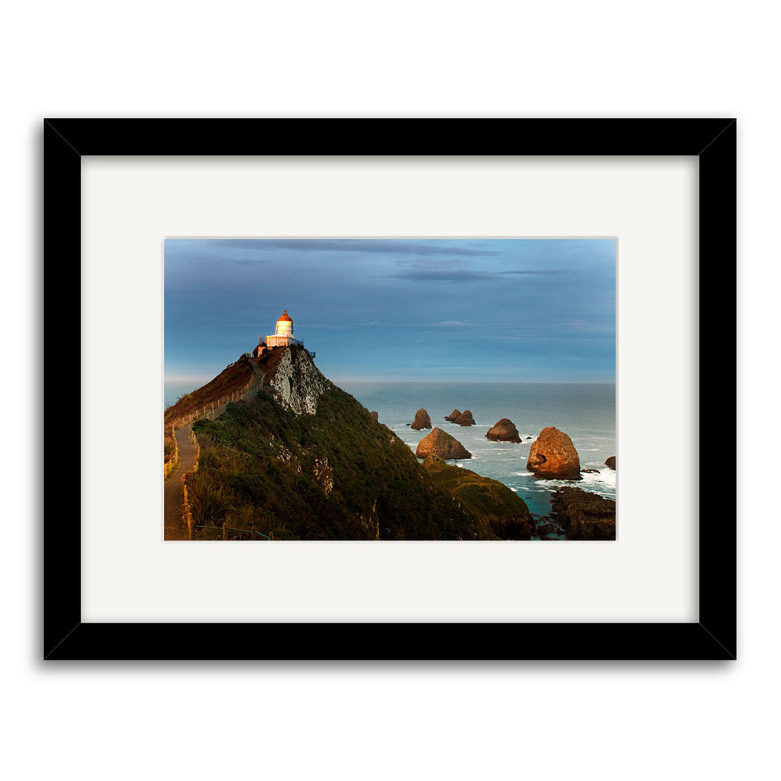 The “Nugget Point Caitlins 2 - RP215” photographic print by Reuben Price shows a lighthouse on a rocky cliff at sunset, with sea stacks below, beautifully framed with a black and white mat on archival-quality paper.
