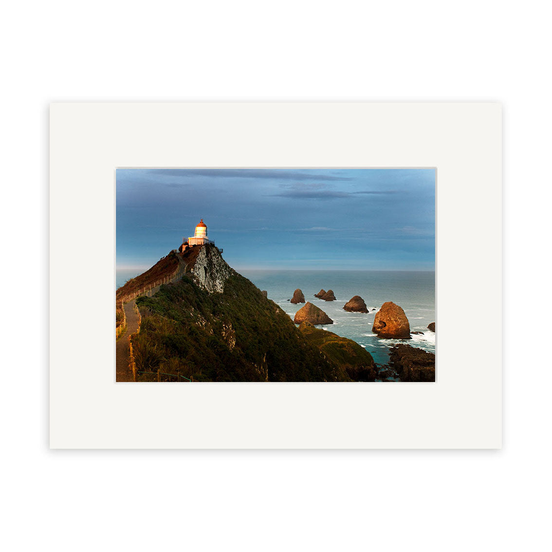 The Nugget Point Caitlins 2 - RP215 print features a lighthouse on a green cliff above the ocean and rocky outcrops, under a cloudy sky. Captured by Reuben Price, this photograph is printed on archival quality paper.