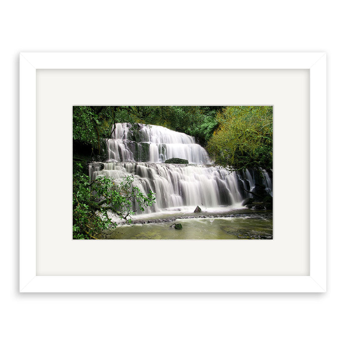 The Purakaunui Caitlins - RP216 is a framed photographic print featuring a multi-tiered New Zealand waterfall amid lush greenery. The white frame and matting accentuate the scene, printed on archival matte paper for enduring beauty.