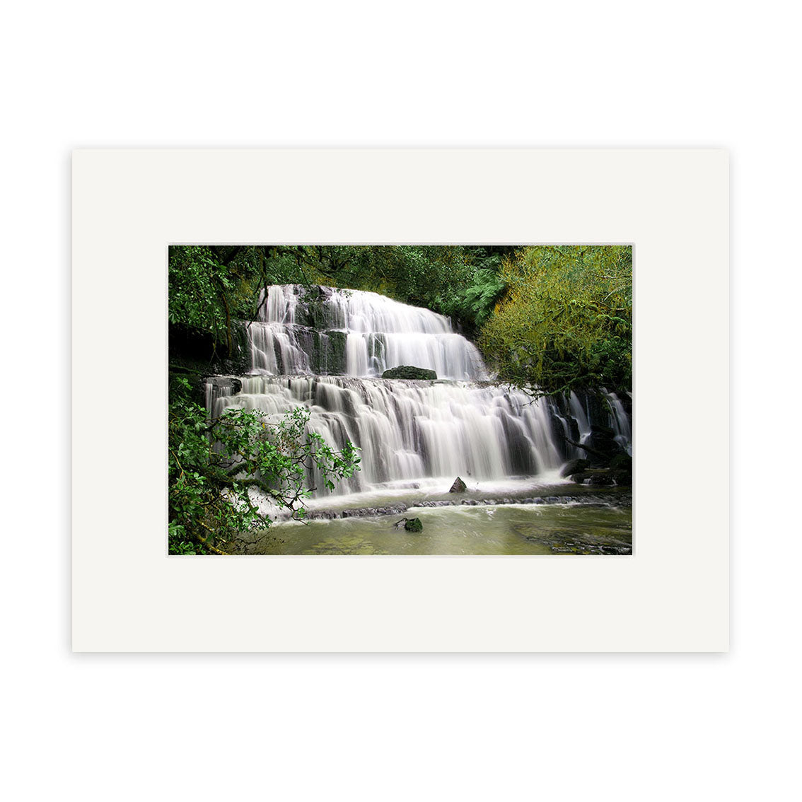 Purakaunui Caitlins - RP216: A photo of a multi-tiered New Zealand waterfall amid lush greenery, with silky water highlighted. Presented in a white mat frame on archival quality matte paper for stunning photographic prints.