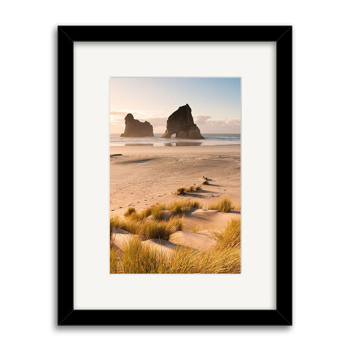 Framed photo Wharariki - RP218 shows a New Zealand beach with tall grasses and two large sea rock formations at sunrise or sunset. Printed on archival matte paper, it comes in a black frame with a white mat.