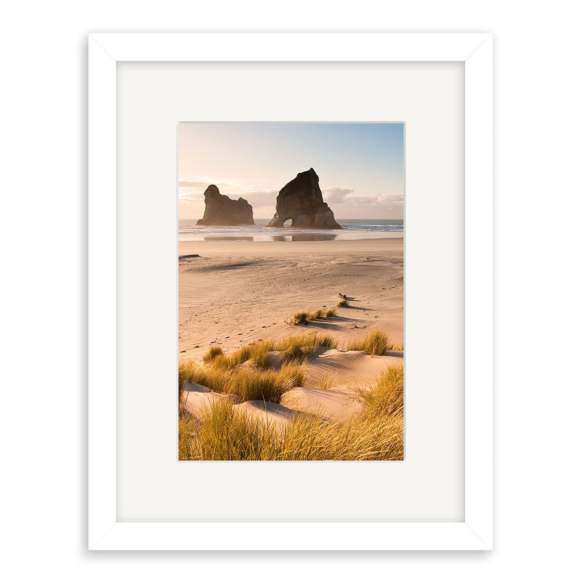 The Wharariki - RP218 is a framed photo of a sandy New Zealand beach with tall grasses, two large rock formations in the ocean, and a partly cloudy sky. A white frame with wide mat enhances the archival matte paper print.