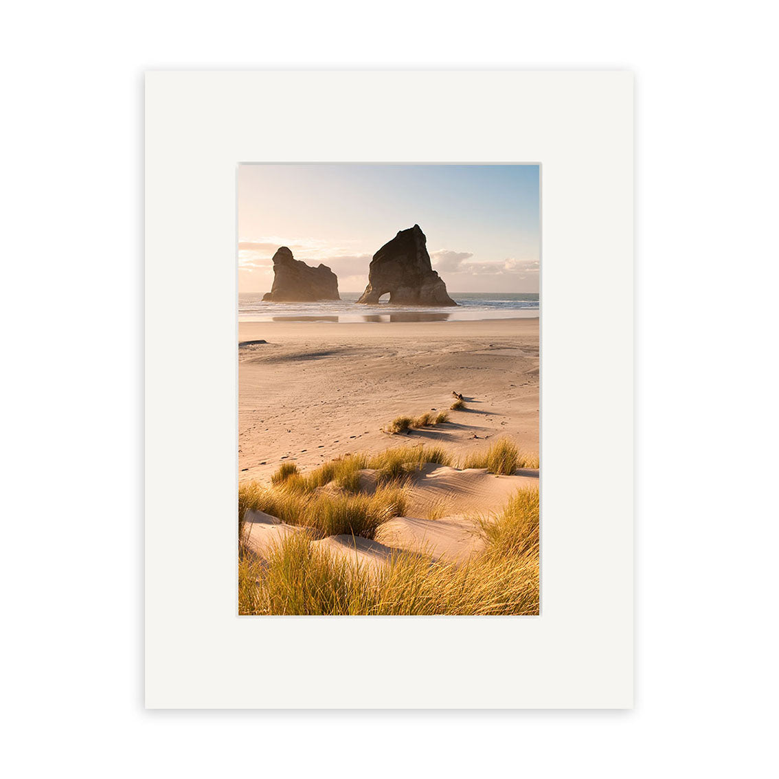 Wharariki - RP218: A photo of a sandy New Zealand beach with tall grasses, two large ocean rock formations, and a partly cloudy sky, displayed in a white mat frame on archival quality matte paper.