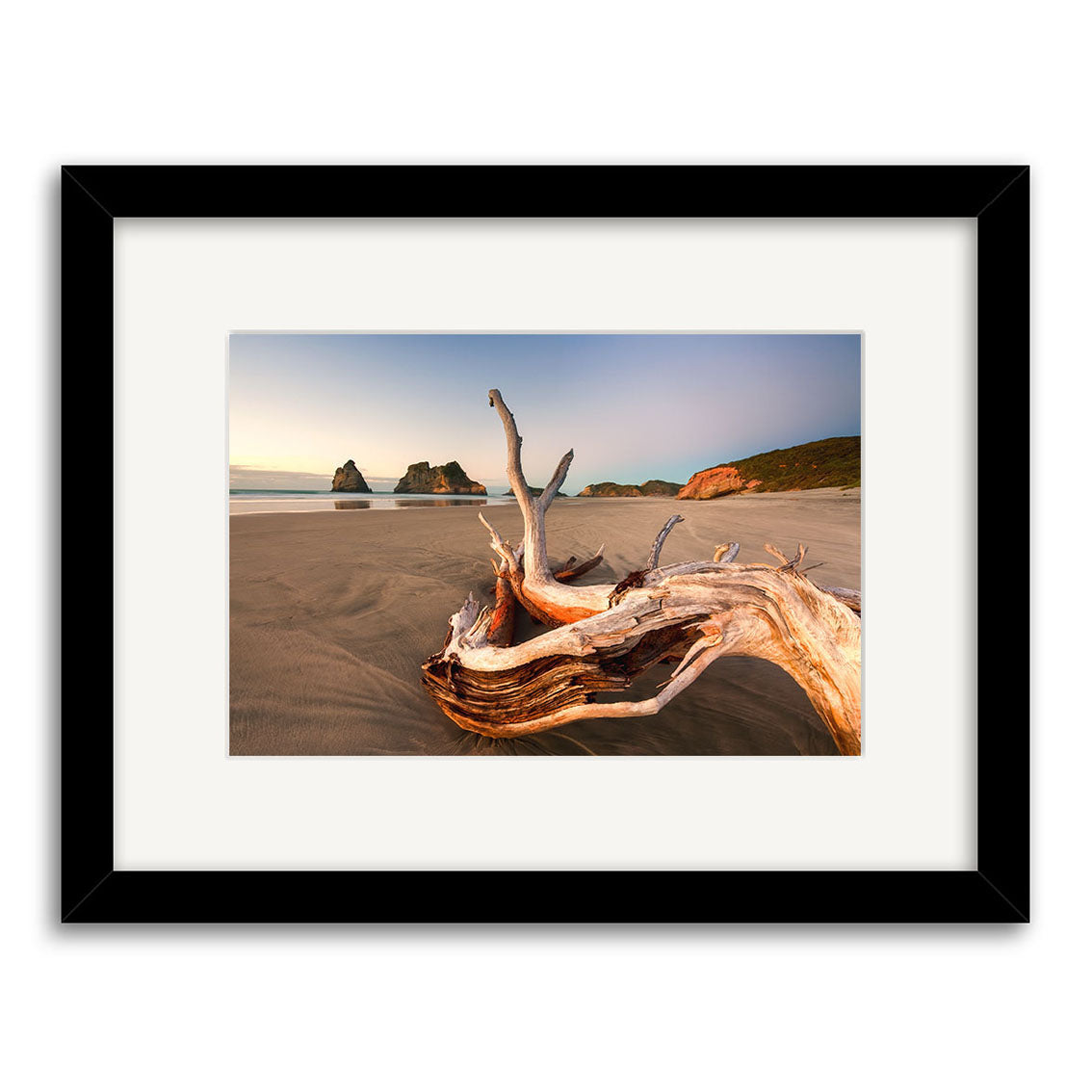 Framed photographic print titled Wharariki 2 - RP219, showcasing a sandy New Zealand beach with driftwood and distant cliffs under a clear sky, printed on archival quality matte paper.