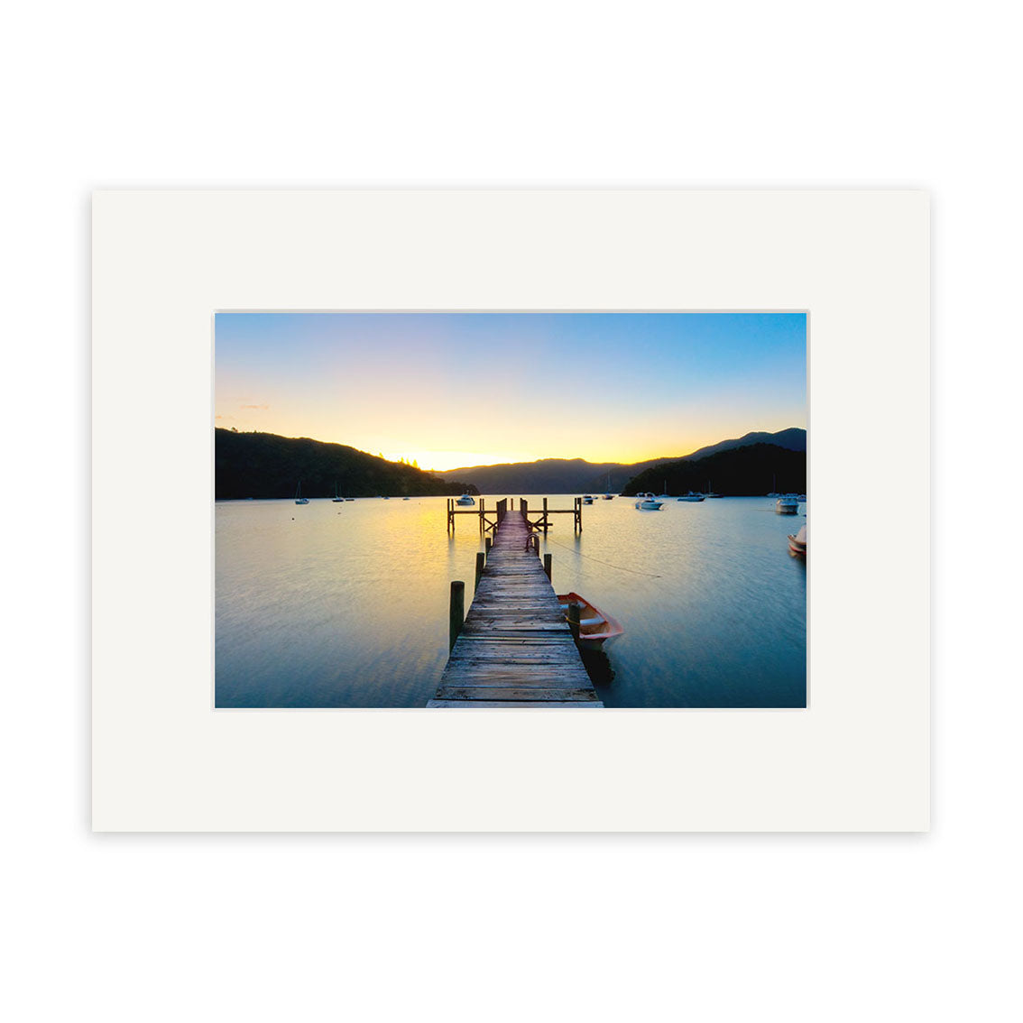 The Marlborough Sounds - RP220 photographic print shows a wooden dock over calm New Zealand waters at sunset, boats anchored nearby, and silhouetted hills. It is matted with a wide white border on archival quality matte paper.