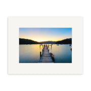 The Marlborough Sounds - RP220 photographic print shows a wooden dock over calm New Zealand waters at sunset, boats anchored nearby, and silhouetted hills. It is matted with a wide white border on archival quality matte paper.