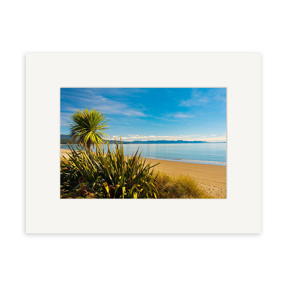 “Golden Bay - RP222” features a sandy beach with green spiky plants, calm blue water, distant mountains, and a blue sky with clouds, presented in a simple white mat frame on archival quality matte paper.