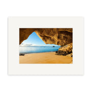 The Kaiteriteri Cave - RP224 print shows a sandy New Zealand beach and calm waves viewed from a rocky cave, with blue sky and clouds. Displayed in a white mat frame on archival-quality matte paper.