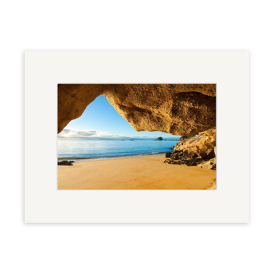 The Kaiteriteri Cave - RP224 print shows a sandy New Zealand beach and calm waves viewed from a rocky cave, with blue sky and clouds. Displayed in a white mat frame on archival-quality matte paper.