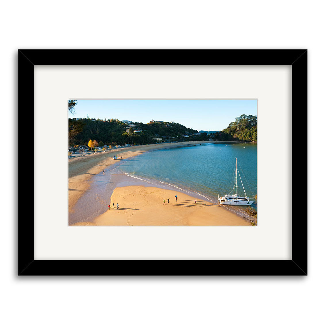 Framed archival quality photographic print, Kaiteriteri - RP225, features a sandy beach with a few people walking, a sailboat on calm blue water, and trees under a clear New Zealand sky.