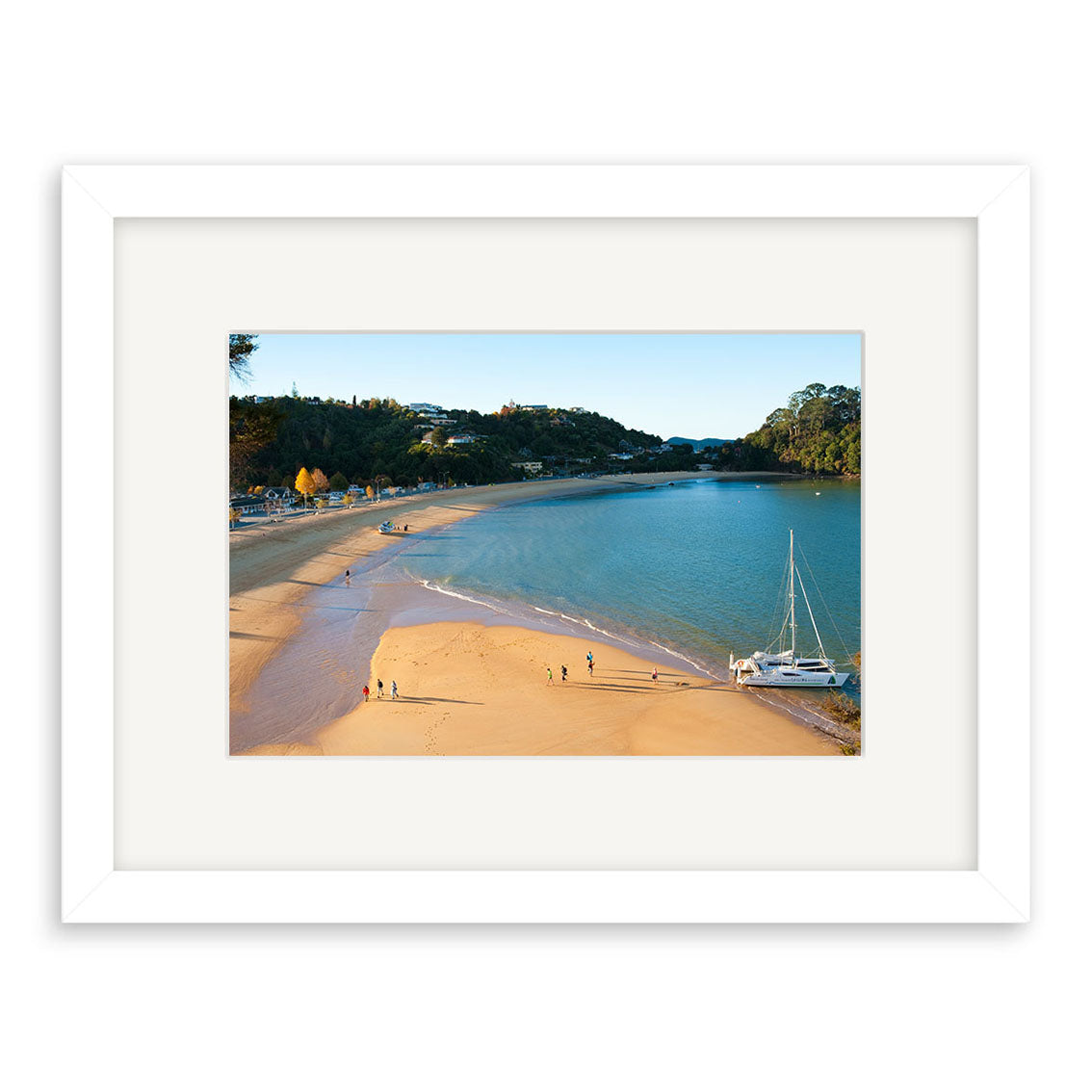 A framed photographic print, Kaiteriteri - RP225, features a New Zealand beach with golden sand, calm blue water, people by the shore, and a white sailboat nearby. Trees and buildings are visible in the background. Archival quality guaranteed.
