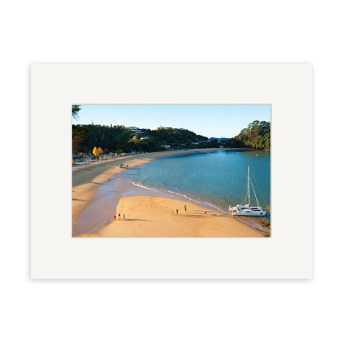 Kaiteriteri - RP225 features a sandy beach curving along a calm New Zealand bay with a sailboat near shore, people walking on the sand, and trees and buildings under a clear sky. The photographic print is framed with a white mat.