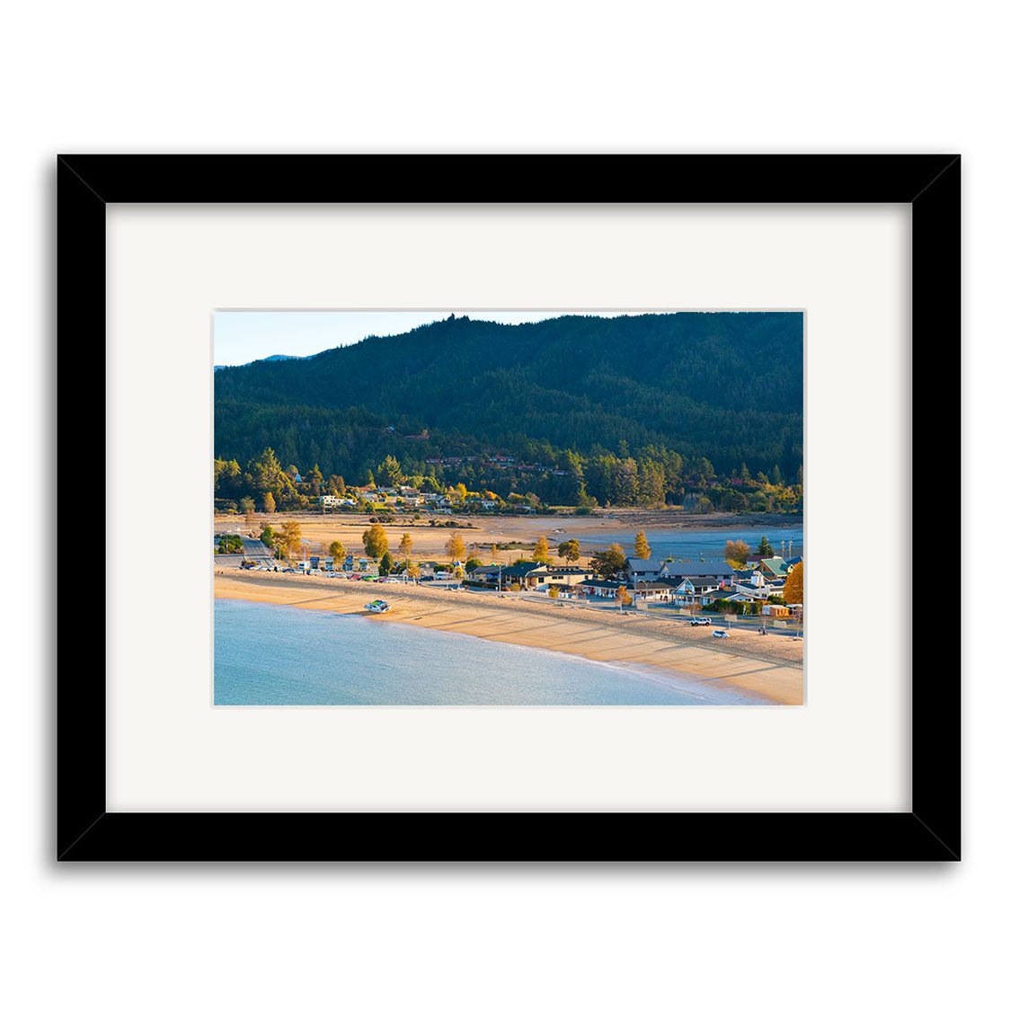 Kaiteriteri 2 - RP226 is a framed photographic print by Reuben Price, depicting a coastal New Zealand town with houses along a sandy beach, water in the foreground, and forested hills behind. Presented with a white mat and black frame.