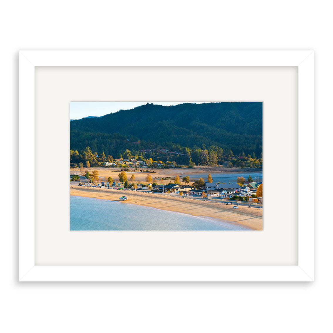 Kaiteriteri 2 - RP226 is a framed photographic print by Reuben Price featuring a sandy New Zealand beach, calm water, shoreline houses, and a forested hill under a clear sky.