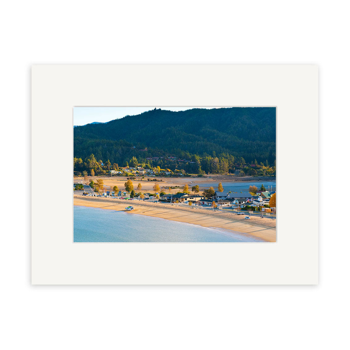 The Kaiteriteri 2 - RP226 photographic print by Reuben Price features a coastal town beside a sandy beach, with houses, trees, and hills in the background under a clear sky, presented in a white mat frame.