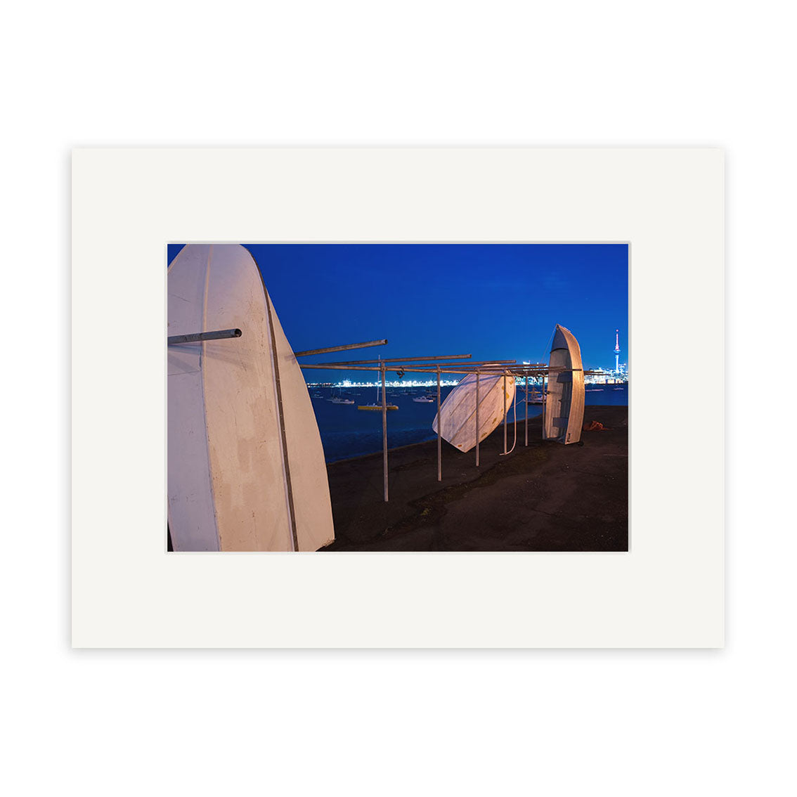 Three small boats are stored upright on a metal rack beside the water at night in Auckland Dinghy - RP228. City lights and a tower appear in the distance under a dark blue sky. This archival quality print comes framed with a wide white mat.