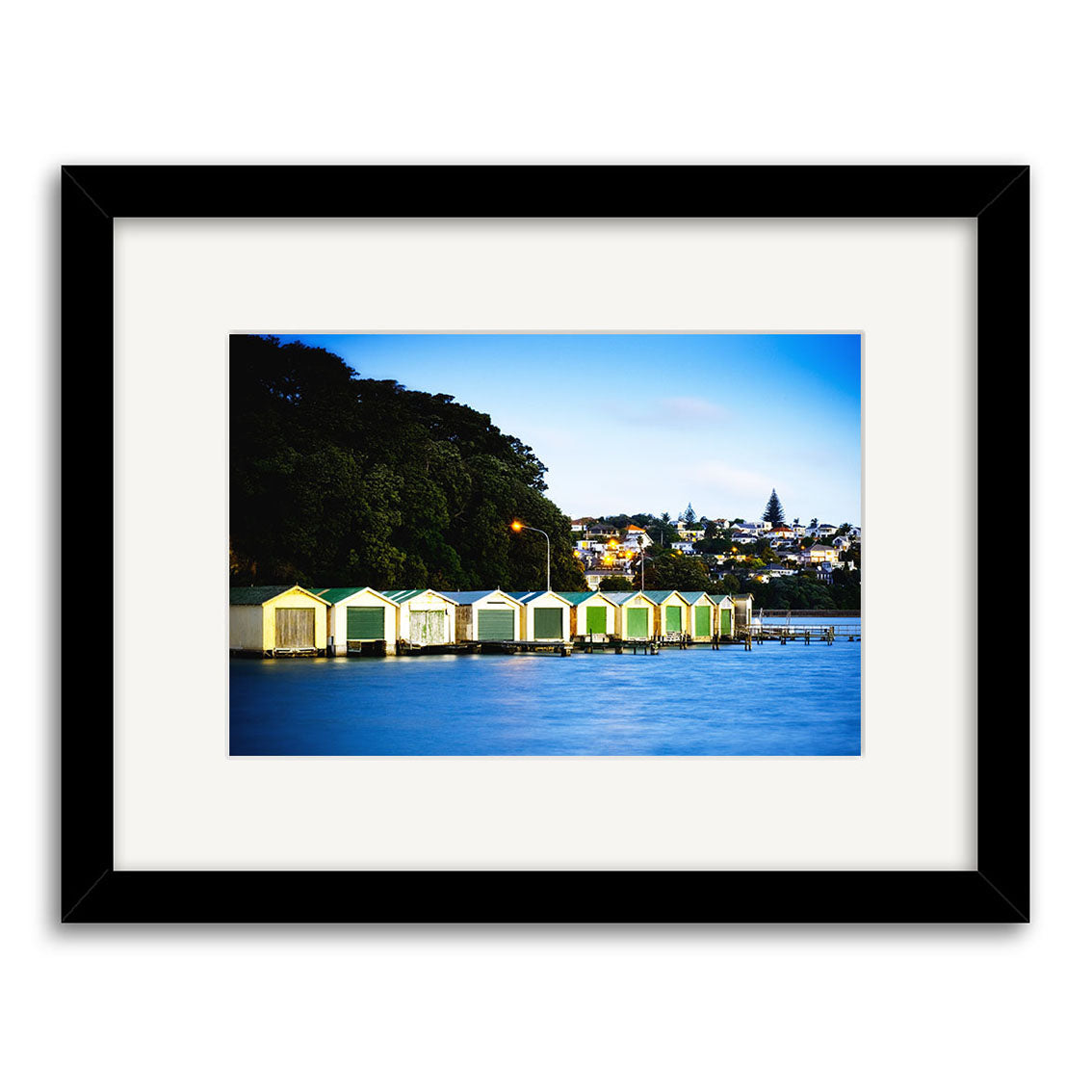 Orakei Boatsheds - RP231: Framed photo of colorful New Zealand boathouses beside calm blue water with hillside homes, trees, and a clear sky—printed as archival quality photographic prints.