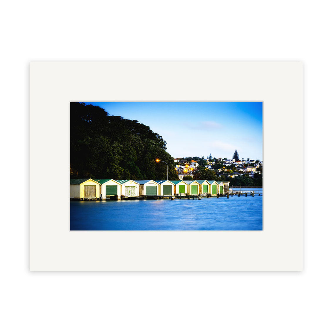 Orakei Boatsheds - RP231 captures a row of vibrant boat sheds by the water under a tree-covered hill in New Zealand, with houses and greenery in the background—ideal for archival quality photographic prints.