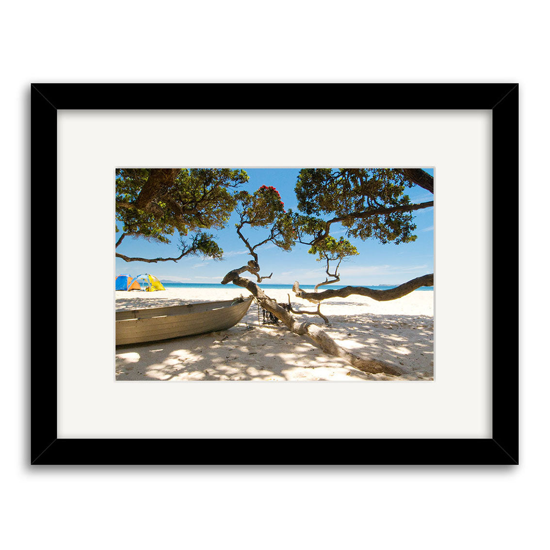 A framed photographic print, Otama Beach - RP233, features a New Zealand beach with a wooden boat on sand, twisting tree branches, blue sky, and a colorful umbrella—printed on archival quality matte paper.