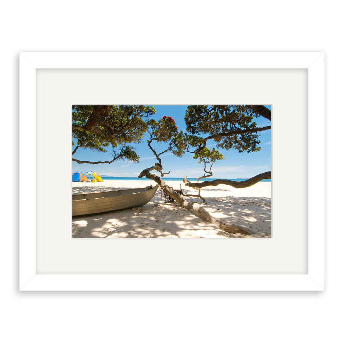 Otama Beach - RP233: A framed photographic print of a sandy New Zealand beach with a small boat, twisted tree branches, blue sky, and beach umbrellas. Matted on archival-quality matte paper in a white frame.
