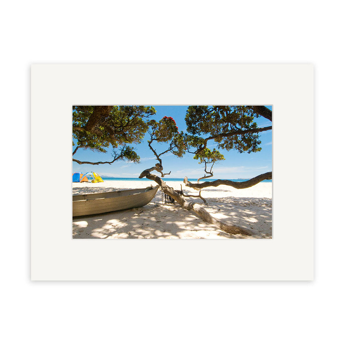 Otama Beach - RP233 features a sandy New Zealand beach with a large tree casting shadows, a wooden rowboat to the left, and colorful umbrellas. The clear blue sky meets the water—ideal for archival quality matte photographic prints.
