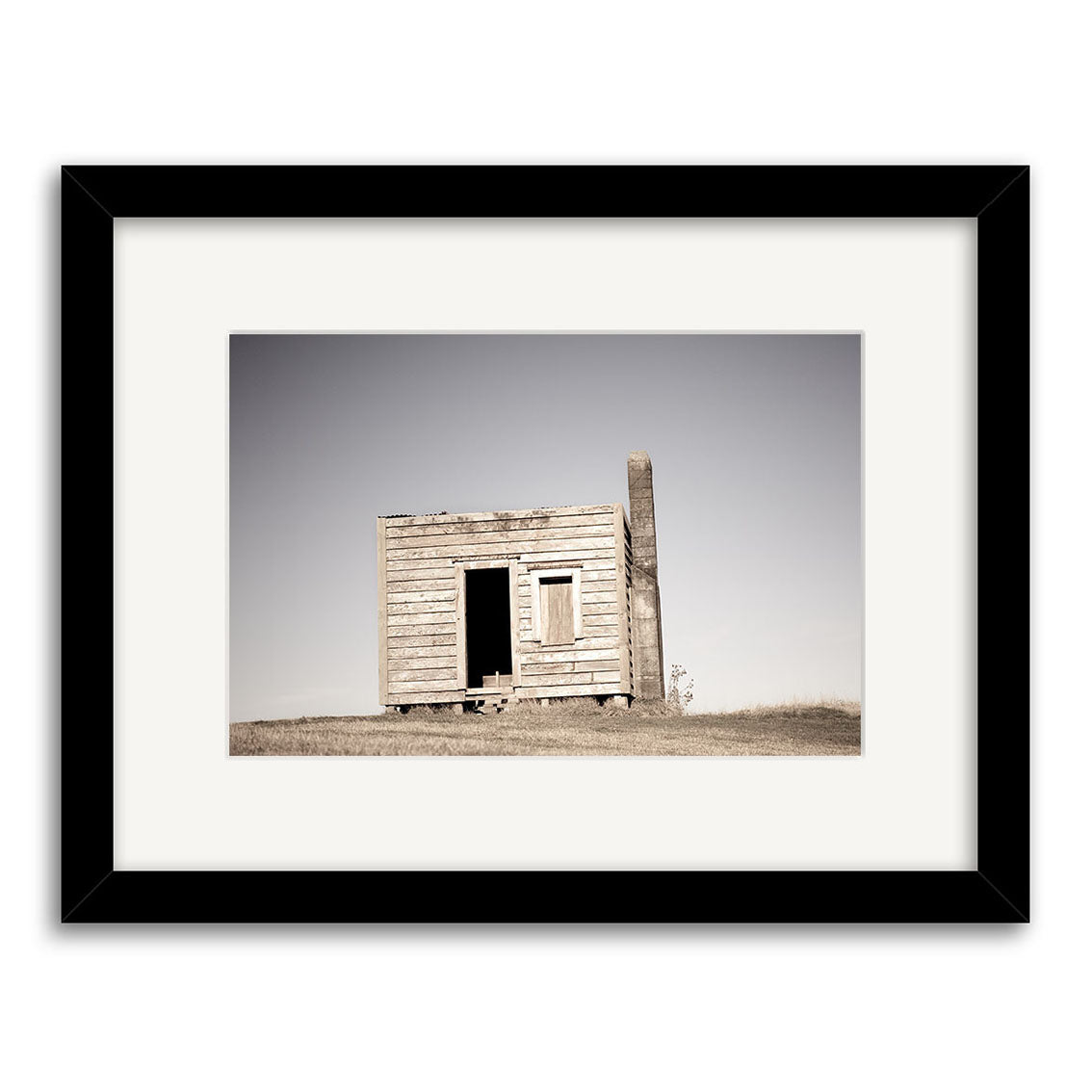 Farm Hut Mamaku - RP236: An archival quality Reuben Price framed photography print showcases a weathered wooden shack with a slanted roof on grassland under clear skies, featuring a muted palette and minimal detail—perfect for New Zealand print collectors.
