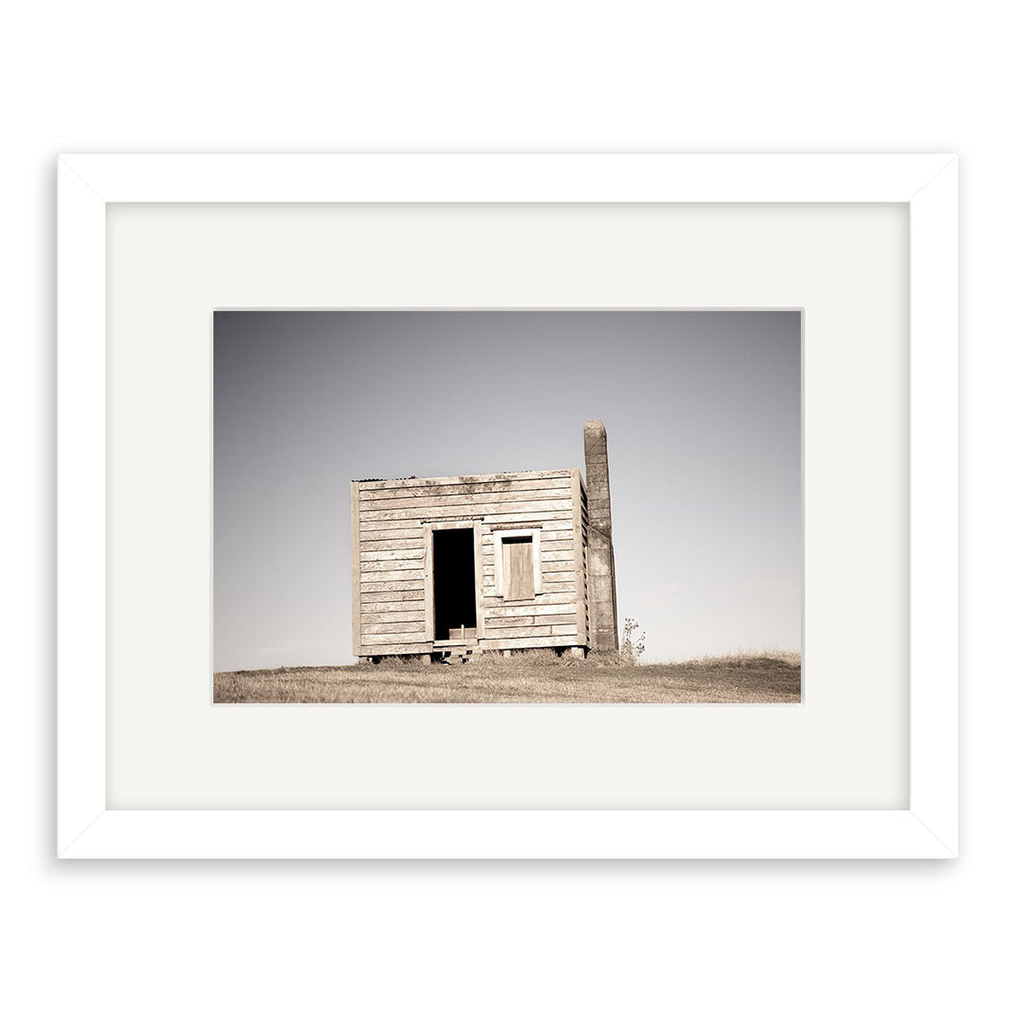 Displayed in a white frame with a white mat, Farm Hut Mamaku - RP236 showcases Reuben Prices photography—a striking image of a small weathered shack with a tilted roof standing alone on a grassy field under clear sky.