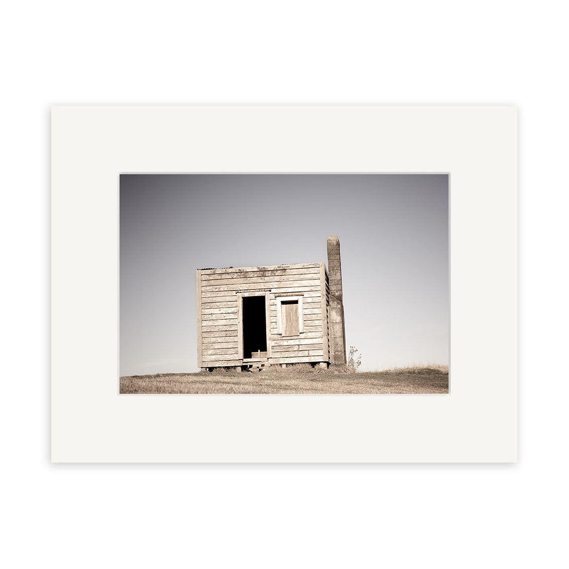 Farm Hut Mamaku - RP236 features a small, weathered wooden shack with a slanted roof in a grassy field under clear skies, capturing the timeless charm of New Zealand in Reuben Prices photographic print.
