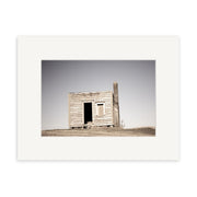 Farm Hut Mamaku - RP236 features a small, weathered wooden shack with a slanted roof in a grassy field under clear skies, capturing the timeless charm of New Zealand in Reuben Prices photographic print.