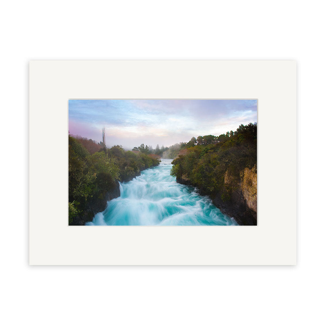 A stunning New Zealand scene, Huka Falls - RP238, showcases white rapids rushing between green, tree-lined banks under a misty sunrise or sunset sky. Presented on archival quality paper with a wide white mat.