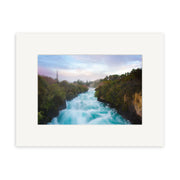 A stunning New Zealand scene, Huka Falls - RP238, showcases white rapids rushing between green, tree-lined banks under a misty sunrise or sunset sky. Presented on archival quality paper with a wide white mat.