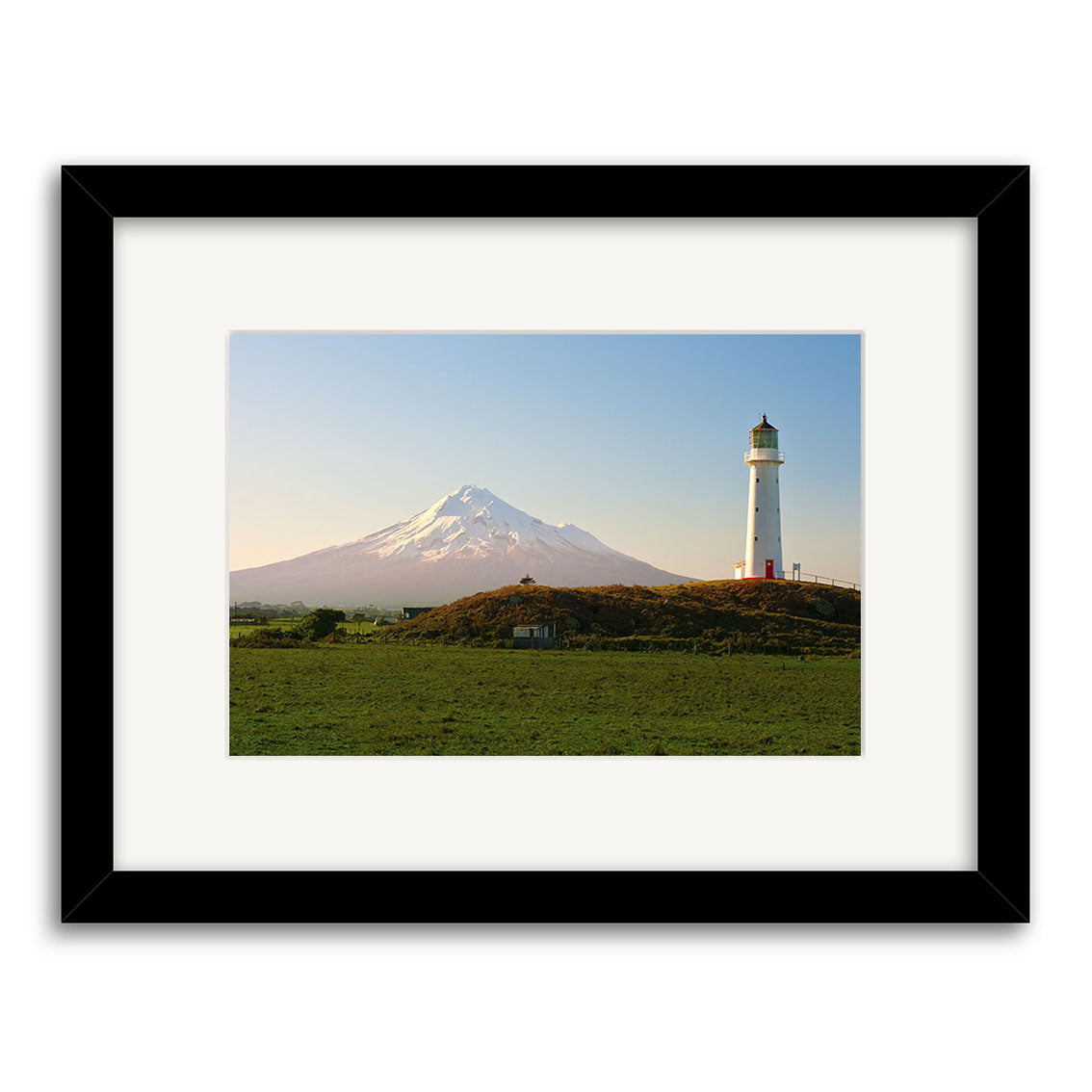 The Cape Egmont - RP240 is a framed photographic print featuring a white lighthouse on a grassy hill in New Zealand, with a snow-capped mountain and clear sky. Printed on archival matte paper for lasting beauty.