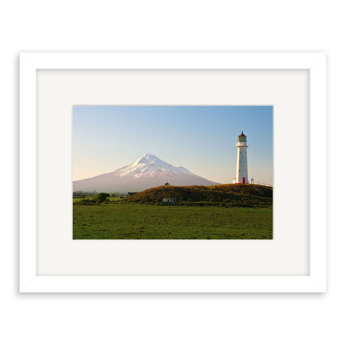 Cape Egmont - RP240: A white lighthouse on a grassy hill with a snow-capped mountain under clear skies, captured in New Zealand and presented as archival quality matte photographic prints in a white frame.