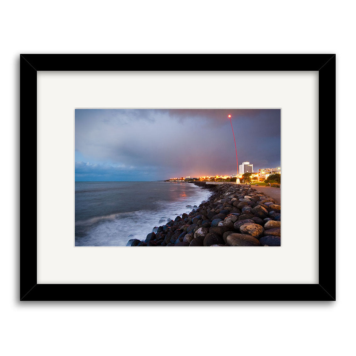 New Plymouth - RP241: Photographic print of a rocky New Zealand shoreline at dusk, with waves, distant city lights, and a red light trail in the cloudy sky. Archival matte paper, black frame.