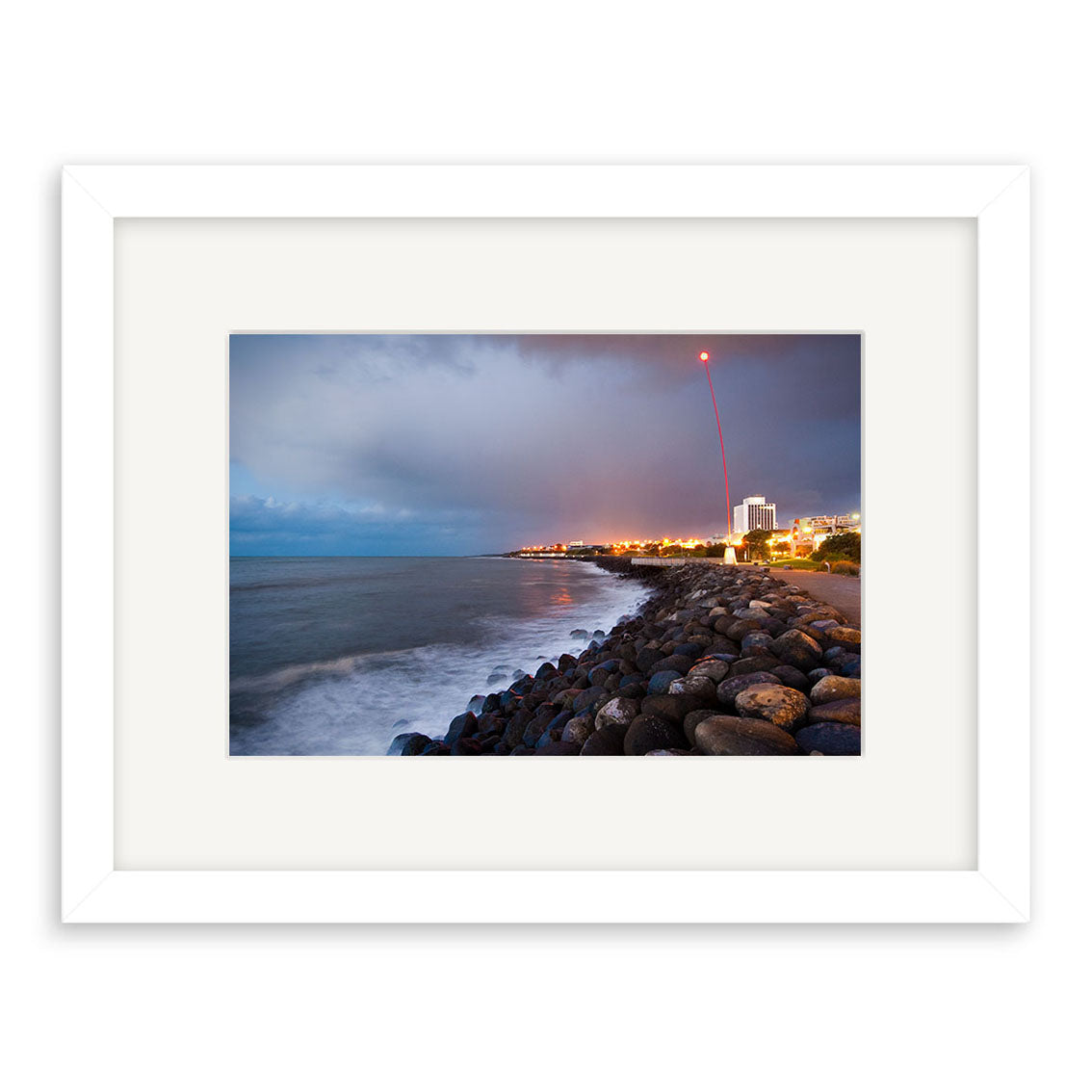 New Plymouth - RP241: A framed photo of a rocky New Zealand coastline at dusk, city lights glowing in the distance, and a red light streak in the cloudy sky. Printed on archival matte paper, gentle waves wash over the shore.
