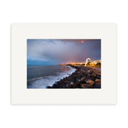 “New Plymouth - RP241” captures a rocky New Zealand shoreline with ocean waves, city lights, and a faint red light trail under clouds. This archival matte photographic print comes beautifully framed with a white mat.