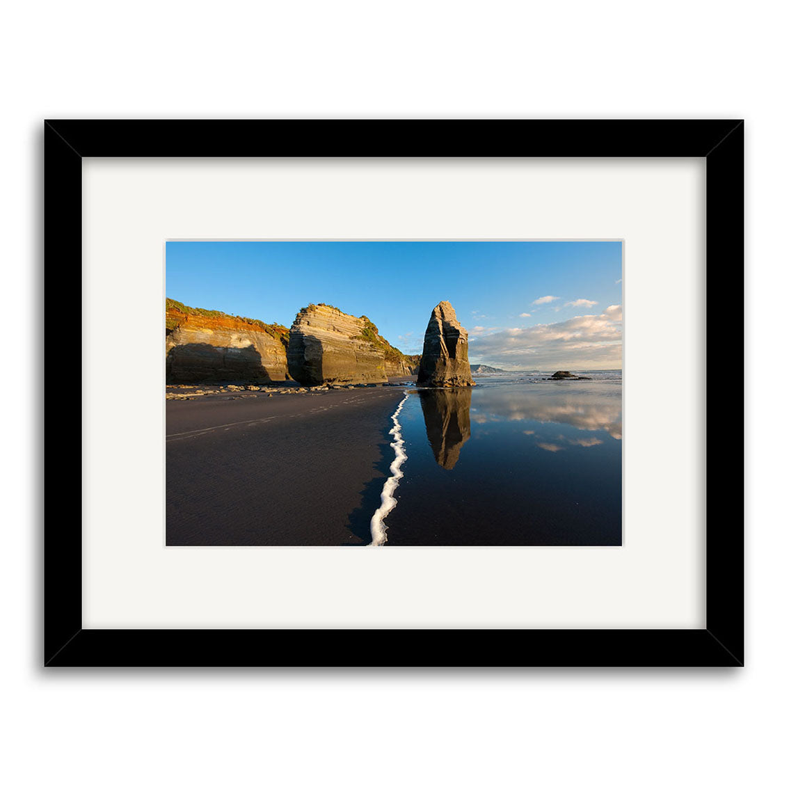 The Tongaporutu - RP242 framed photo print features New Zealand sea stacks reflected in wet black sand, with white foam and a blue sky. Printed on archival quality matte paper for lasting beauty.
