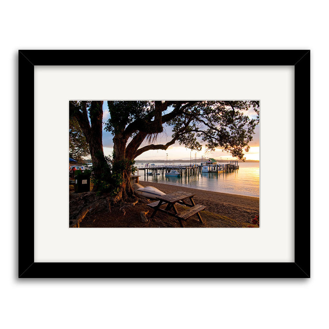 Russell - RP243 is a framed photographic print of a New Zealand waterfront at sunset, featuring a large tree, bench, picnic table, and dock under warm skies. Printed on archival quality matte paper.