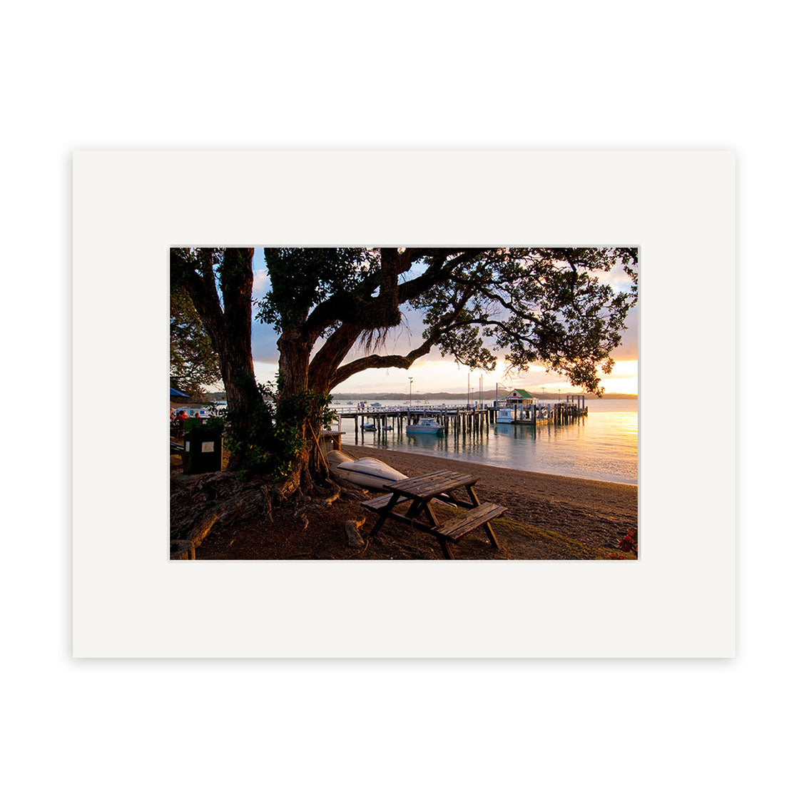 The Russell - RP243 captures a wooden picnic table under a tree by a calm New Zealand waterfront at sunset, with boats at the dock, all viewed through a white photo mat—ideal for photographic prints.