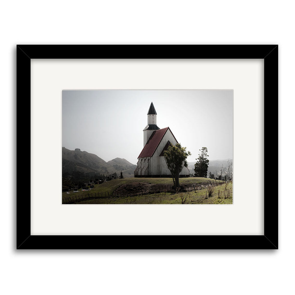 Pawarenga - RP244: A framed archival matte print of a small white church with a red roof and steeple on a grassy New Zealand hill, mountains and trees behind, beneath a cloudy sky.